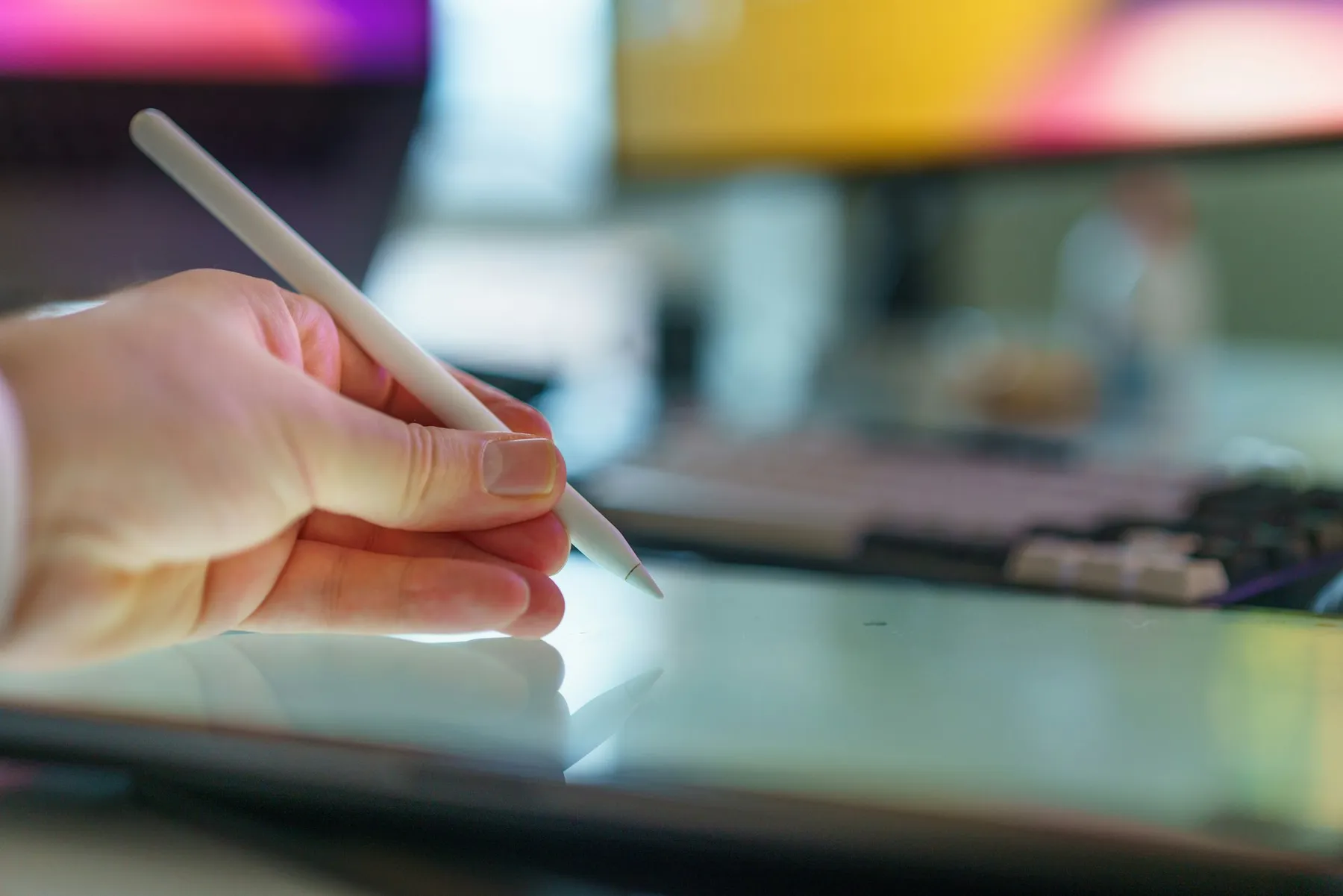 Close-up of a hand holding a digital stylus above a tablet screen, colorful bokeh lighting in the background